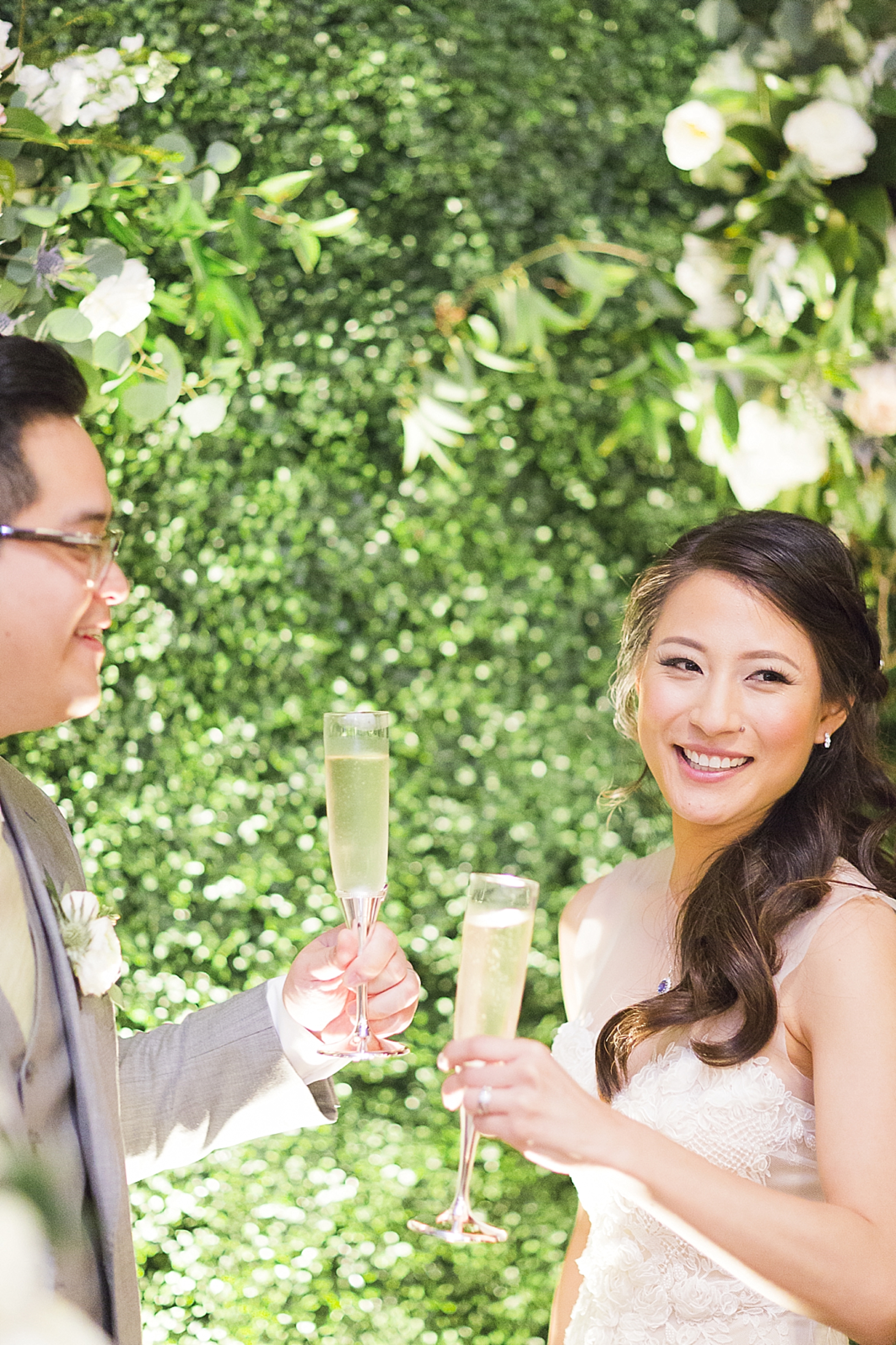 bride and groom champagne in front of wall of greenery
